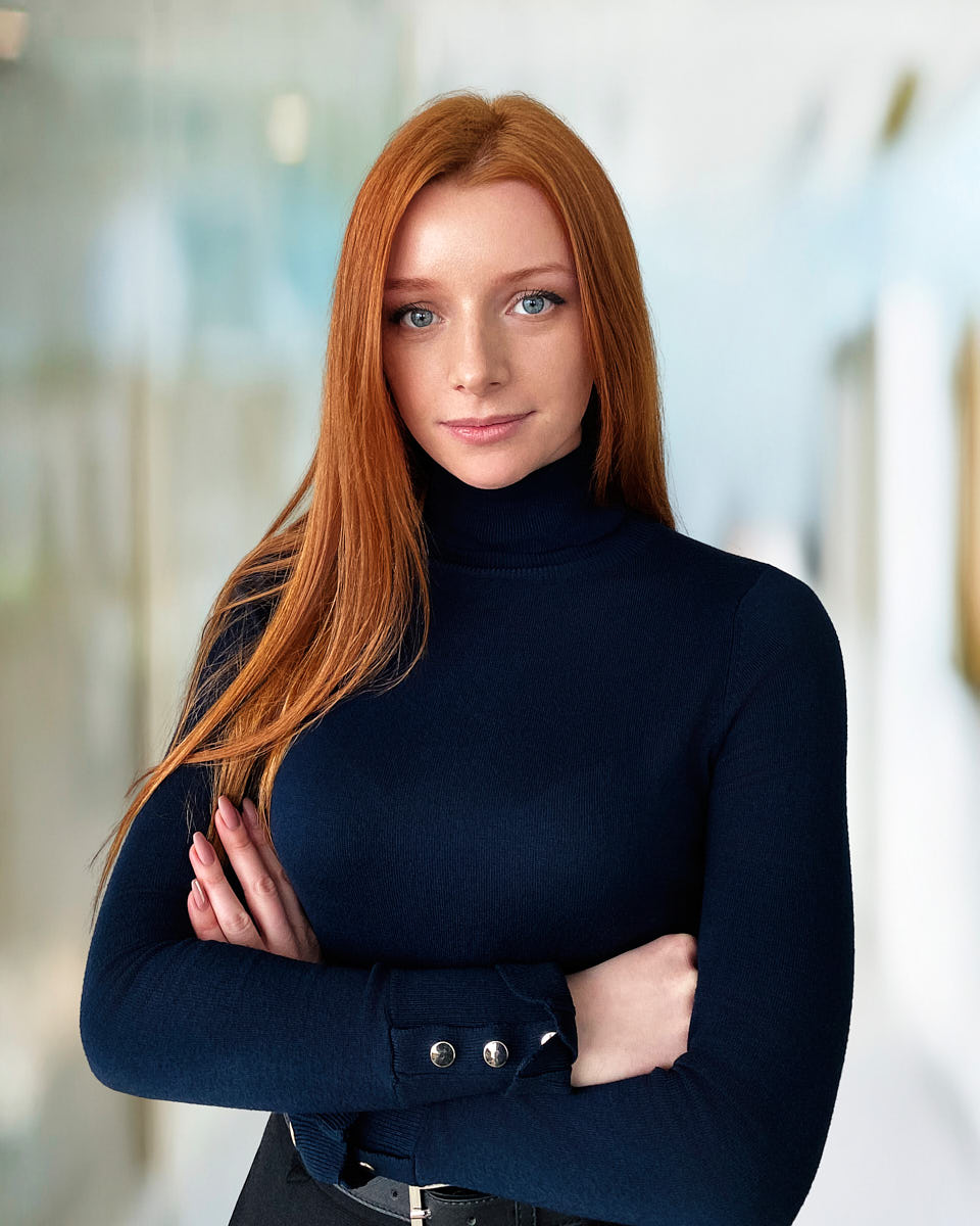 Remote headshot portfolio example of a red-haired woman in a black turtleneck against a bright office background.