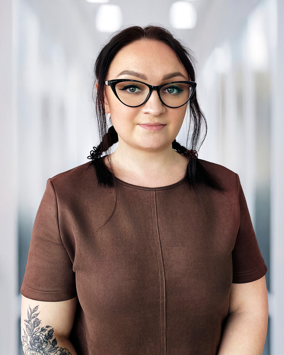 Remote headshot of a woman in glasses photographed against a soft office hallway background.