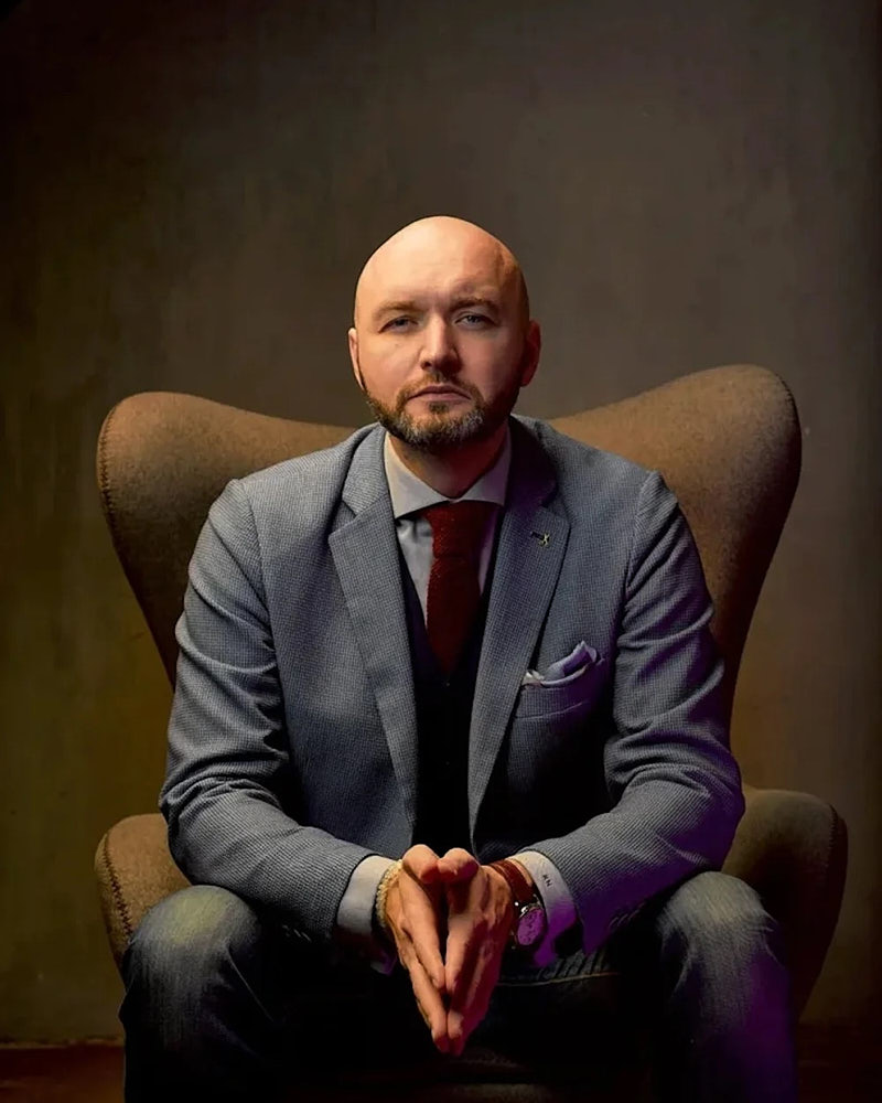 Personal branding portrait of a bald man in a suit seated in a chair against a dark warm background.