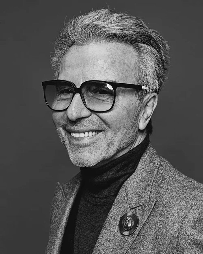 Black and white professional portrait of a person in glasses and a blazer against a clean studio background.