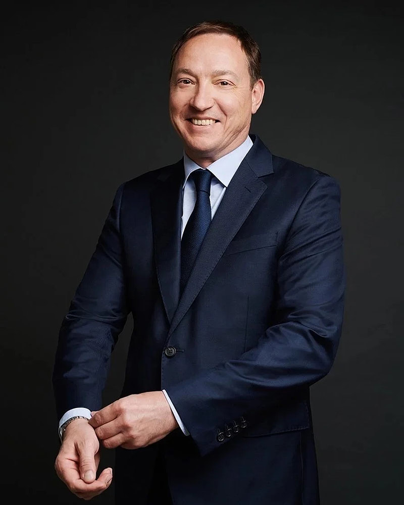 Professional headshot of a man in a navy suit photographed with controlled studio lighting on a dark background.