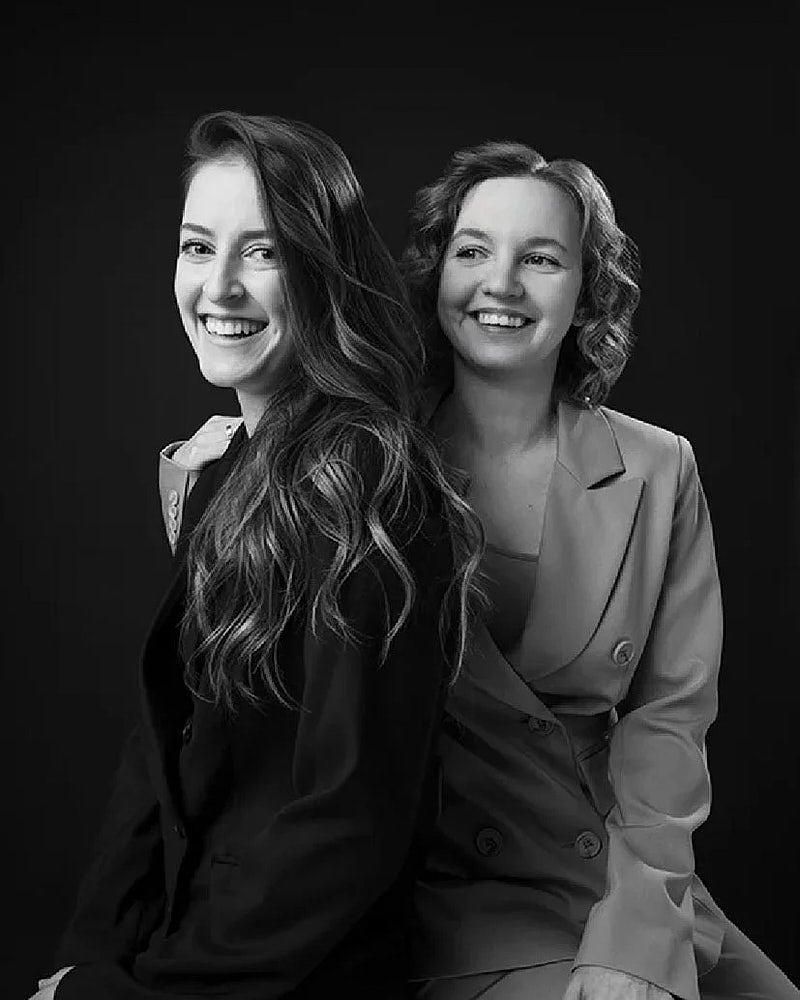 Studio portrait of two women photographed together in a clean black and white professional style.
