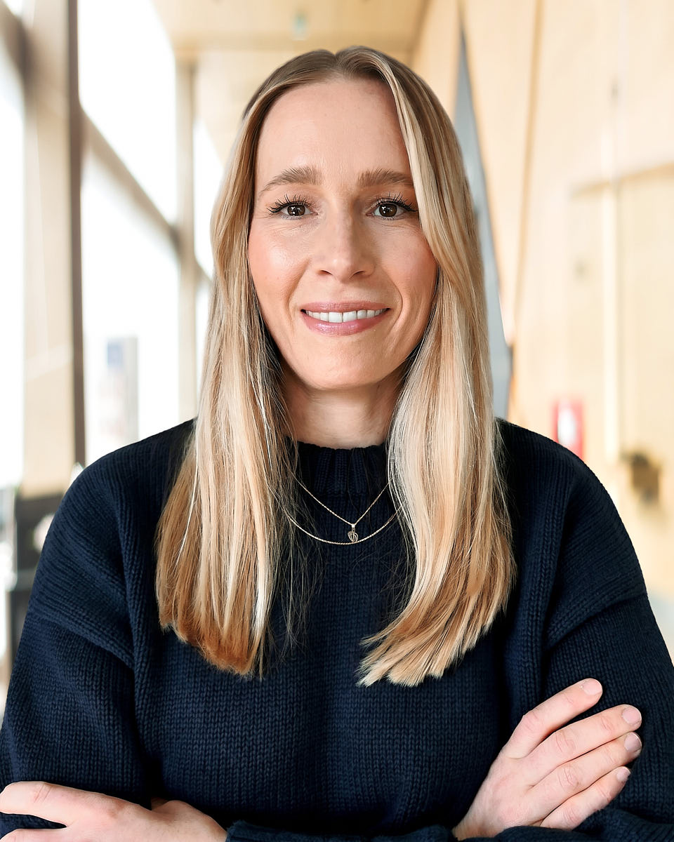 Remote headshot of a blonde woman in a black top photographed in a bright office setting.