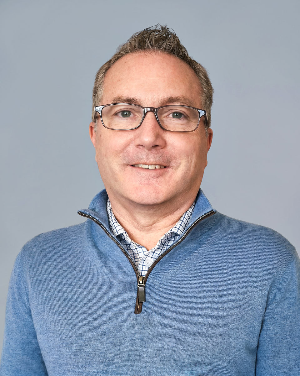 Remote headshot of a man in a blue sweater photographed against a soft gray background.
