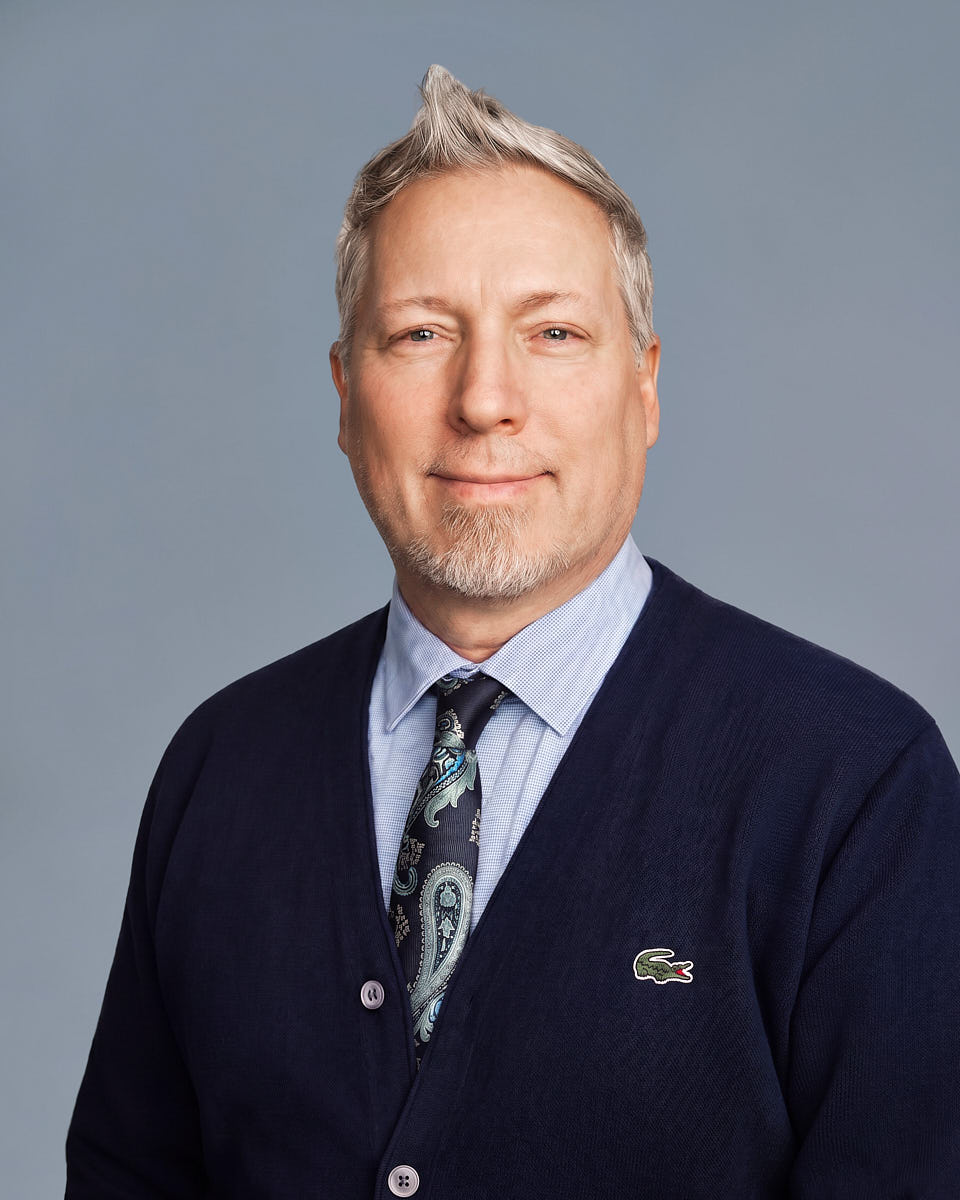 Remote headshot of a man in a dark suit photographed against a blue-gray studio-style background.