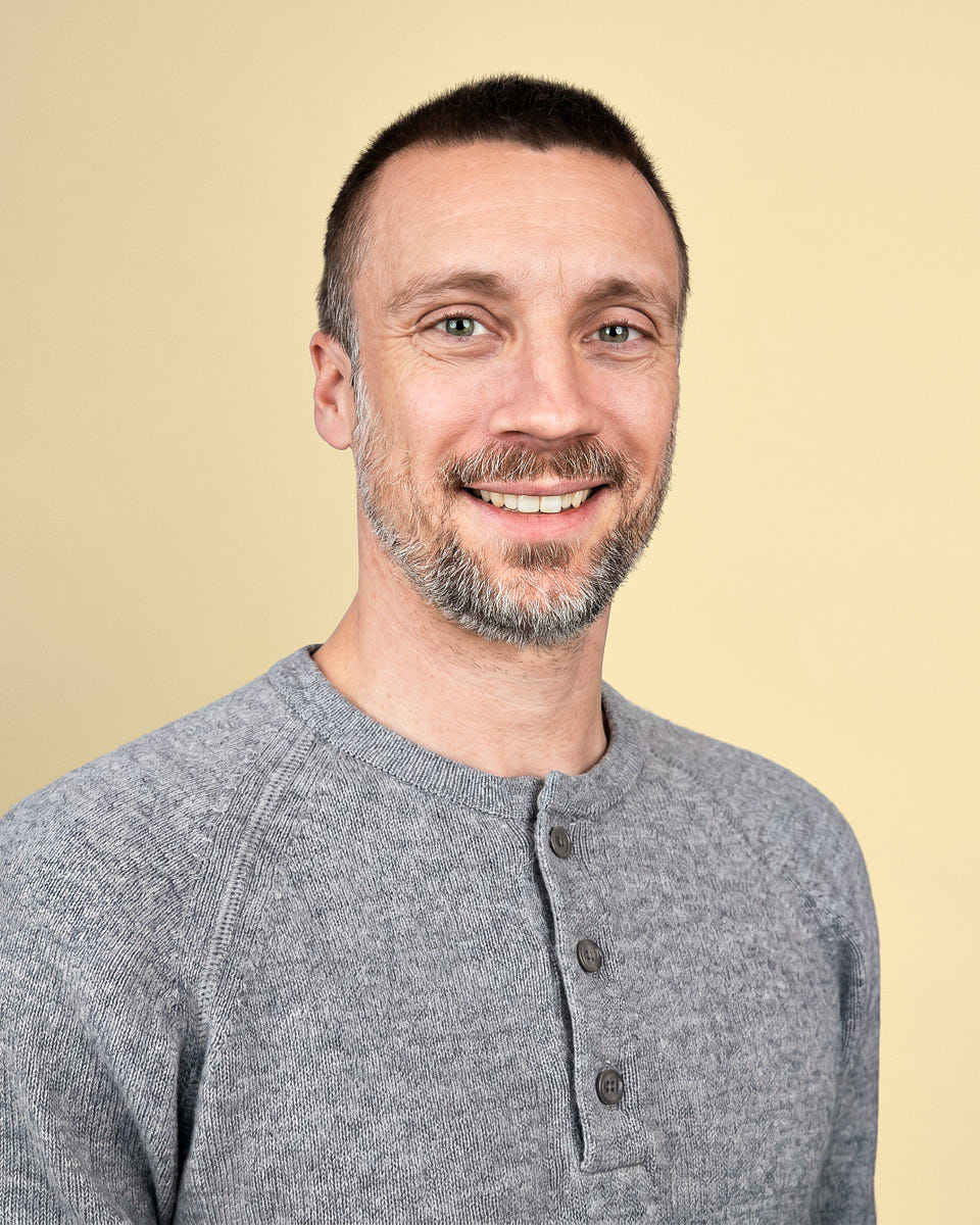 Remote headshot of a man in a gray henley shirt photographed against a warm yellow background.