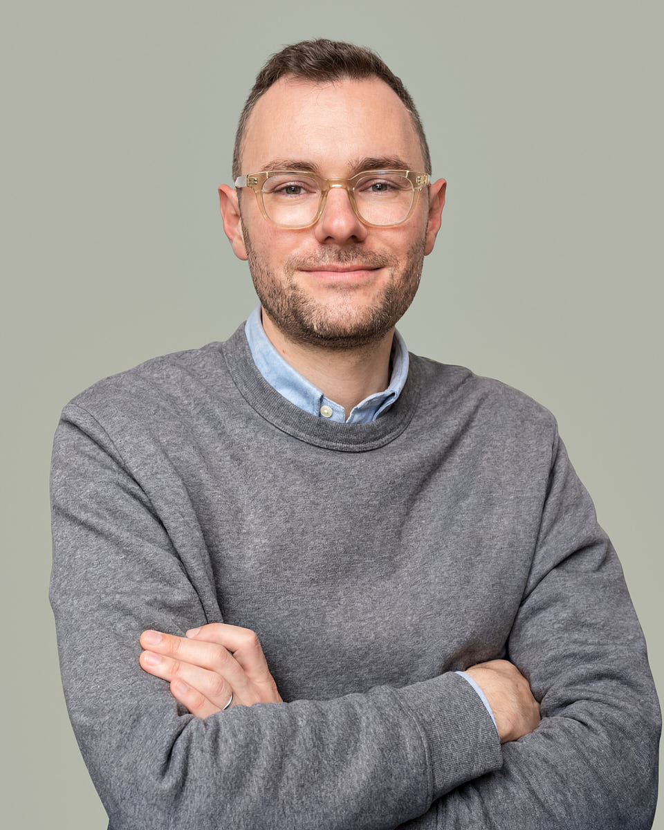 Remote headshot of a man in a gray sweater with arms crossed against a muted green background.