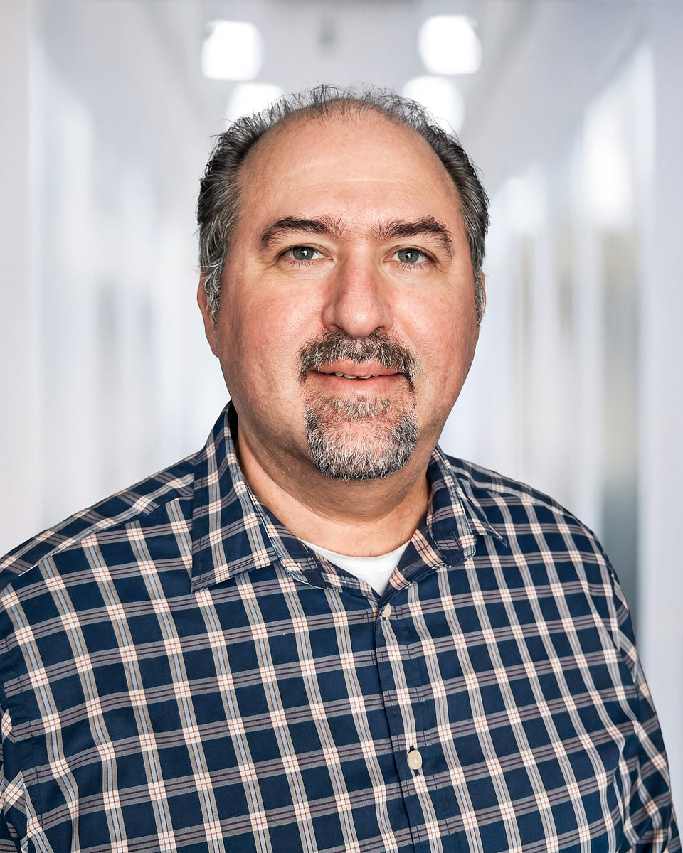 Remote headshot of a man in a plaid shirt photographed against an office hallway background.