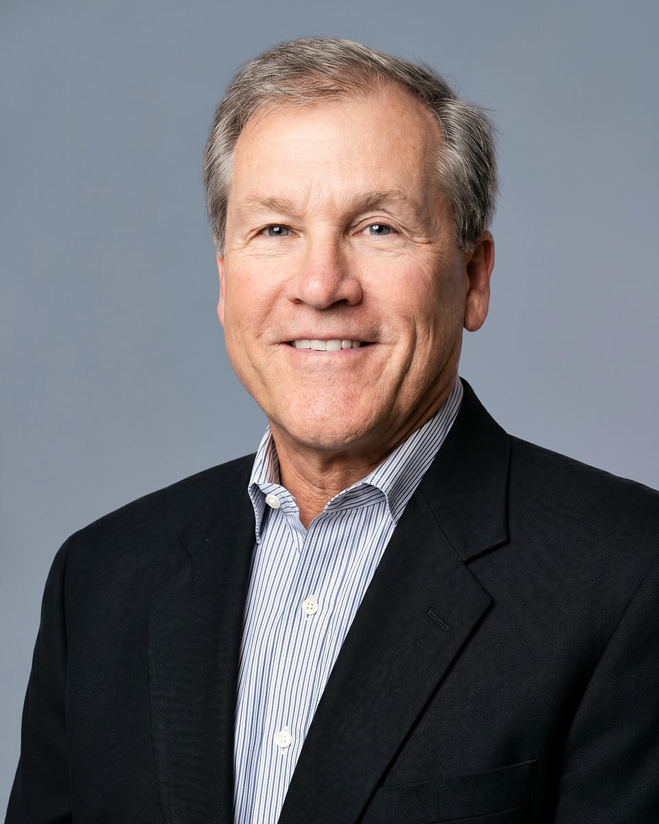 Remote headshot of an older man in a dark blazer photographed on a gray studio-style background.