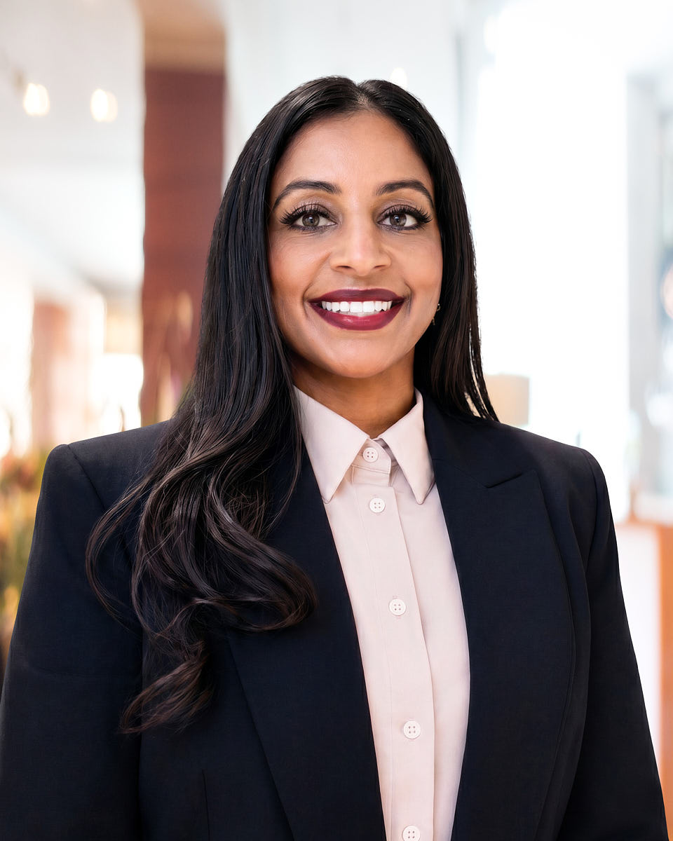 Remote headshot of a woman in a black blazer and light pink shirt photographed against an office background.