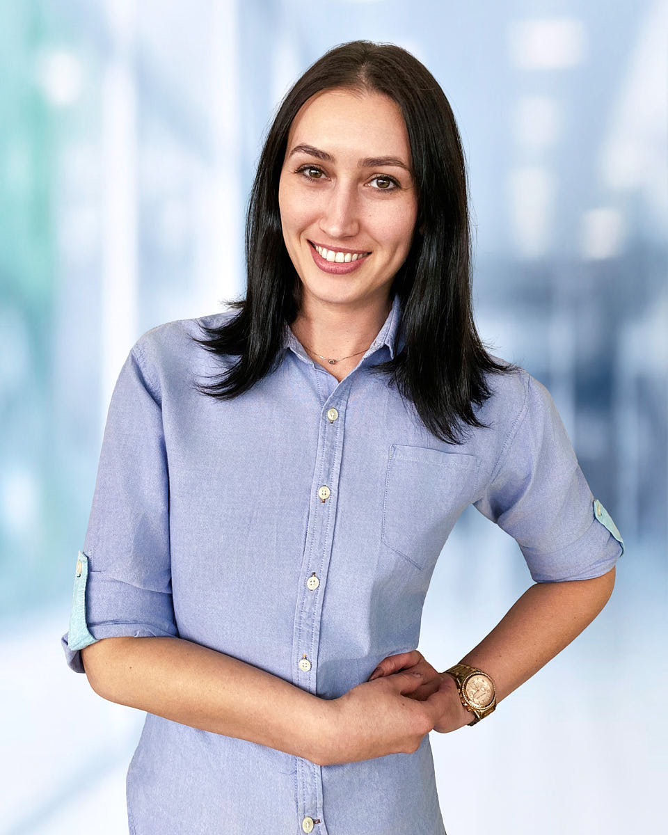 Remote headshot of a woman in a blue button-up shirt photographed against a bright office background.