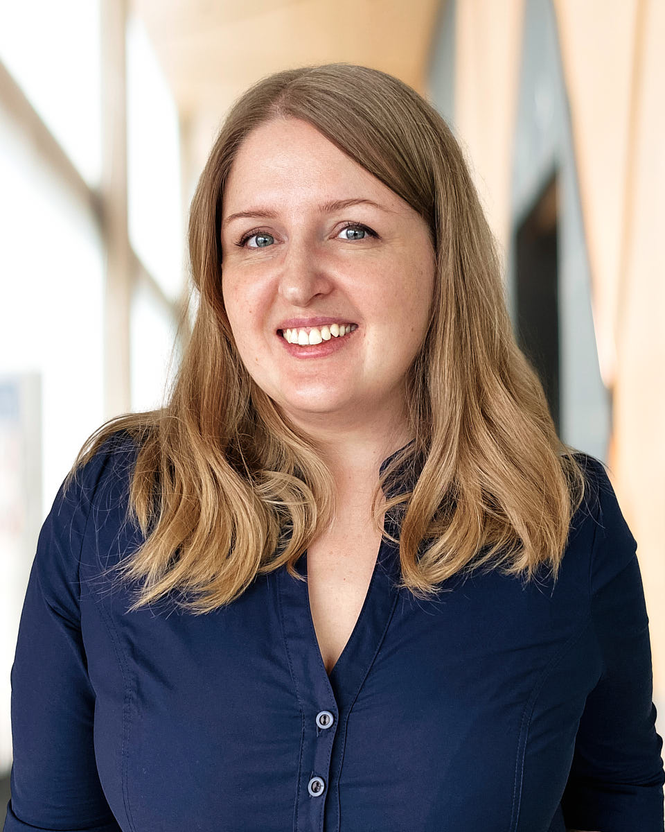 Remote headshot of a woman in a blue shirt photographed with a natural smile in a bright office setting.