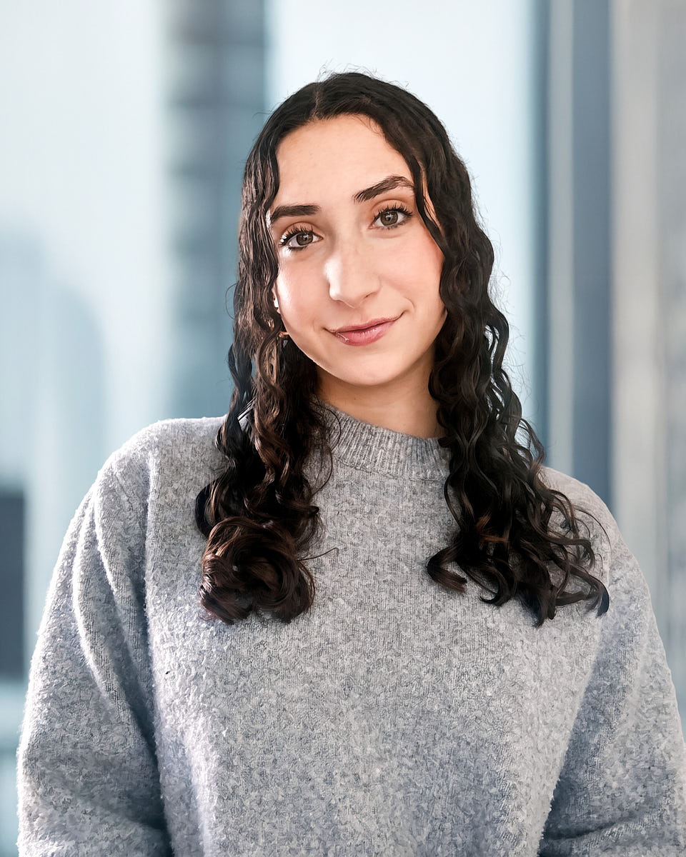 Remote headshot of a woman in a gray sweater photographed against a softly blurred office background.