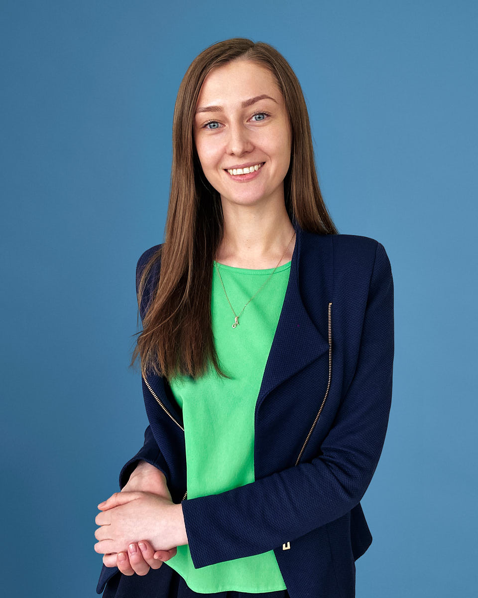 Remote headshot of a woman in a green shirt and navy blazer photographed on a blue studio-style background.