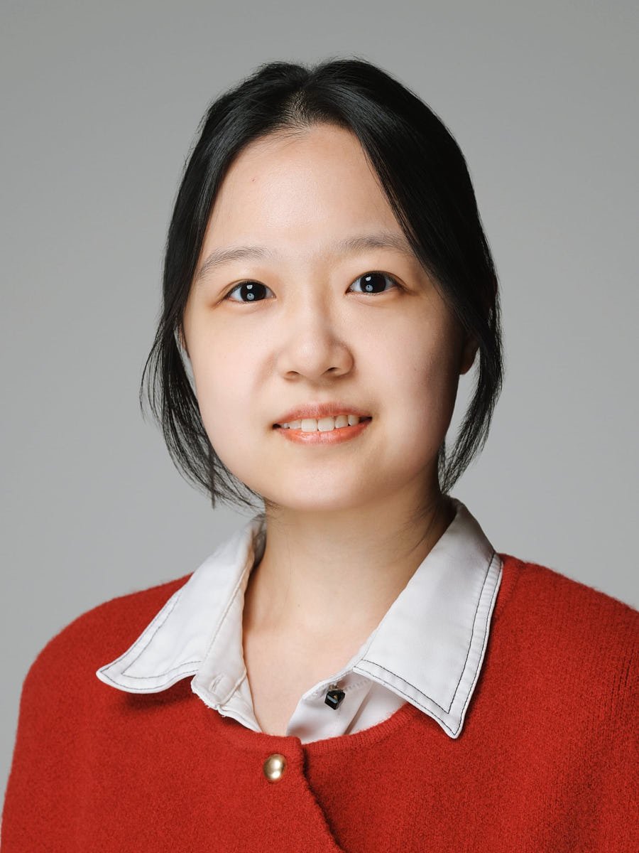 Studio headshot of an Asian woman in a red sweater and white collar against a gray background.