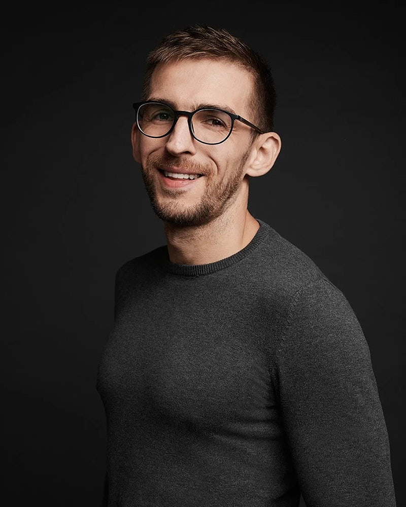 Team headshot of a man in glasses photographed against a dark studio background.